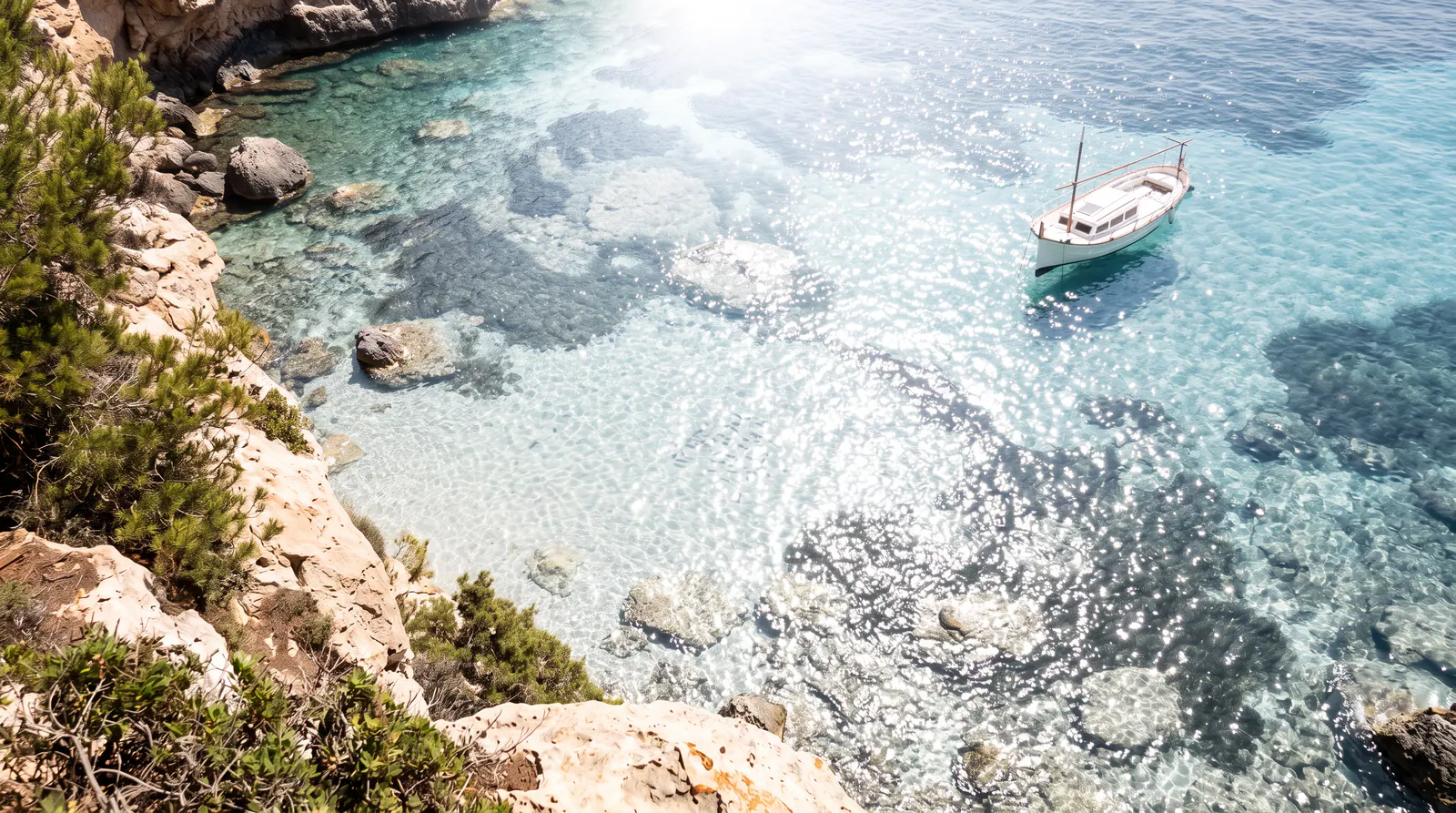 Cala de Mallorca vista desde el acantilado a mediodía: el sol hace brillar el agua y no deja ver el fondo. Con lente polarizada aparecen la arena, la posidonia, las rocas y un banco de sargos.